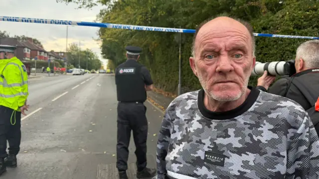 Steven has short shaved hair and is wearing a shirt with grey-scale camouflage print. The Police tape is behind him, and there's a photographer taking a picture, and some police officers in the background.