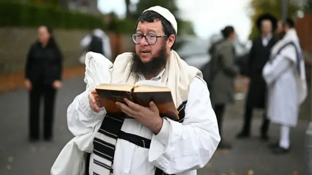 A Jewish man holds a holy book while dressed in white near the Manchester synagogue