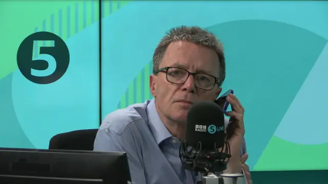 Nicky Campbell takes a call on his phone-in show. He is wearing a blue shirt and glasses and is holding a phone up to the microphone.