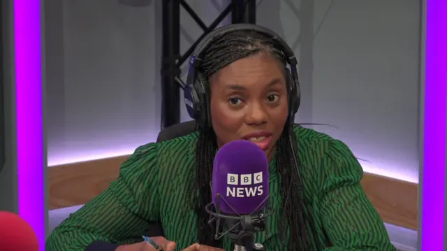 Kemi Badenoch pictured in a BBC studio with a green shirt and a purple microphone in front of her.