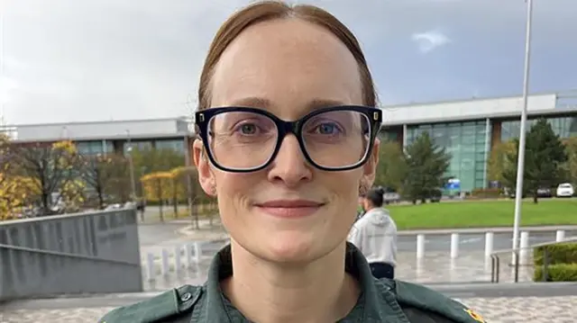 Caroline Lloyd with brown hair in a pony tail wearing black rimmed glasses and her green North West Ambulance Service shirt. She is smiling.