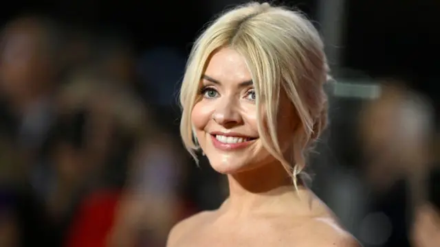 Holly Willoughby is wearing a shoulderless dress. She has blonde hair tied back and is smiling.
