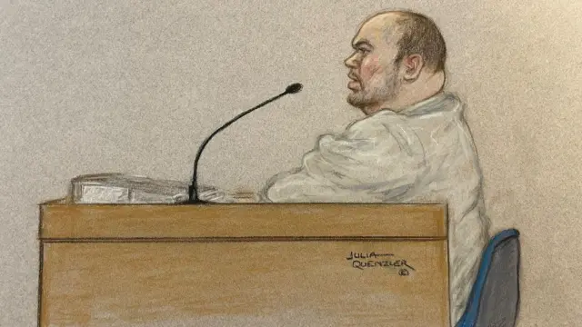 A court sketch of Gavin Plumb giving evidence at court. He has a brown desk in front of him and a black microphone. He is sitting on a blue chair, wearing a grey sweater and talking into the microphone.