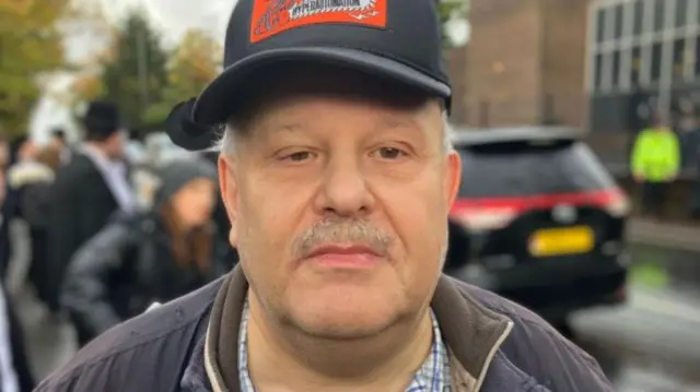 Alan Rubins wearing a black baseball cap and navy jacket with a grey moustache standing outside the synagogue