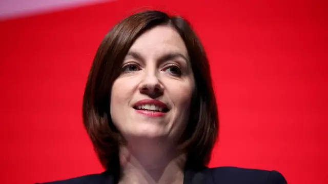 Bridget Phillipson close up head and shoulders photograph, she stands in front of a red background