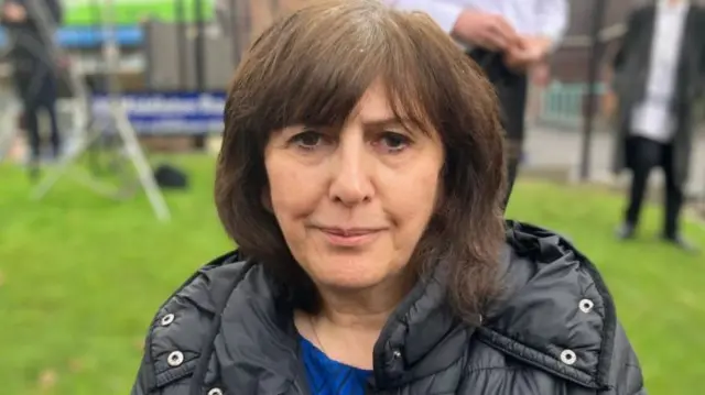 Gloria Levene with shoulder length dark hair, blue jumper and black coat