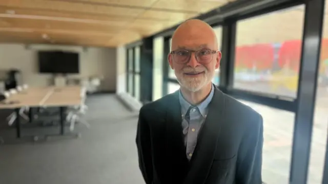 Ian Dunn smiles into the camera, stood in a seminar room at Coventry University. He is a bald man with a white beard and glasses, wearing a dark blazer over a blue shirt.