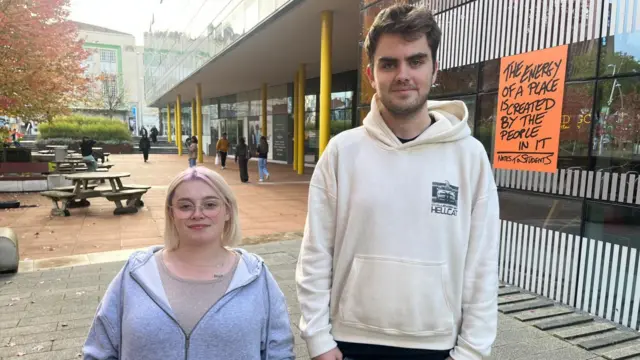 Katie has blonde hair and is wearing a grey hoody and glasses. Toby has brown hair and is wearing a white hoody and jeans