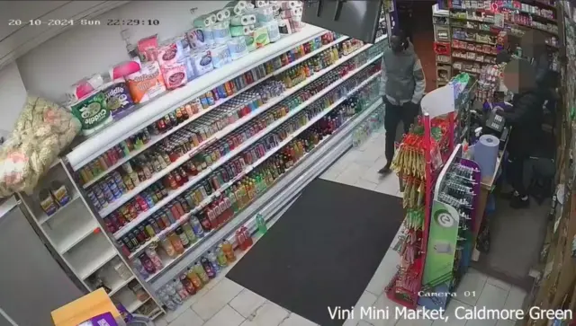 A man in a grey jacket in a small shop with shelves of multi coloured items