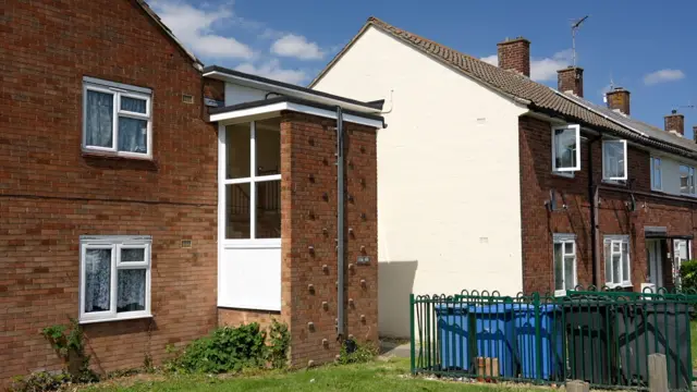 A picture of some flats on a housing estate made of red brick. There is a lawn and some bins at the front and blue sky above.