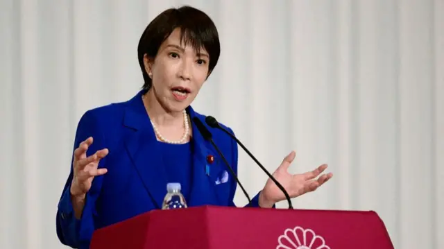 Takaichi, wearing a bright blue suit, gestures with her hands as she speaks at a podium