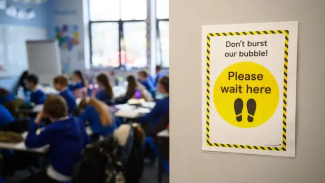 A guidance sign reading 'Don't burst our bubble! Please wait here' can be seen on a door in front of a room full of school pupils sat in a lesson.