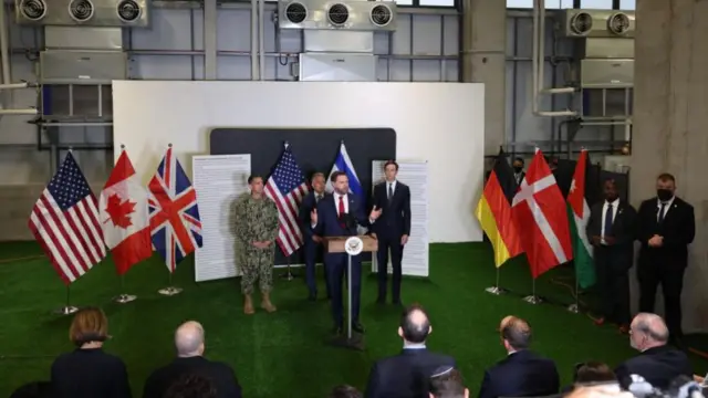 A wide shot showing Vance standing at a podium on a green floor with Witkoff and Kushner behind him