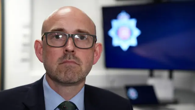 Greg Wood has stubble and is wearing glasses, a black suit jacket, dark green tie and a pale blue shirt. Behind him is the Essex Police crest on a computer monitor