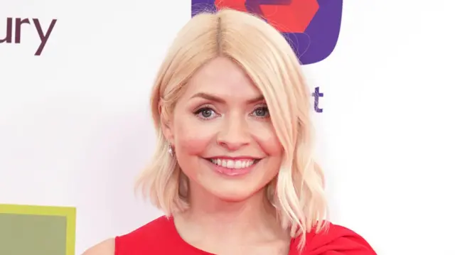TV presenter Holly Willoughby wears her hair in a wavy bob aka a WOB and is wearing a red dress. She is standing in front of a white board with lots of different business logos on