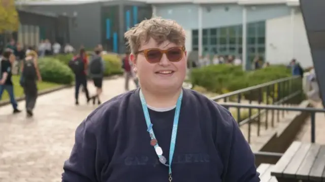Teenager wearing sunglasses, a jumper, and a student lanyard with badges pinned onto it smiles for camera