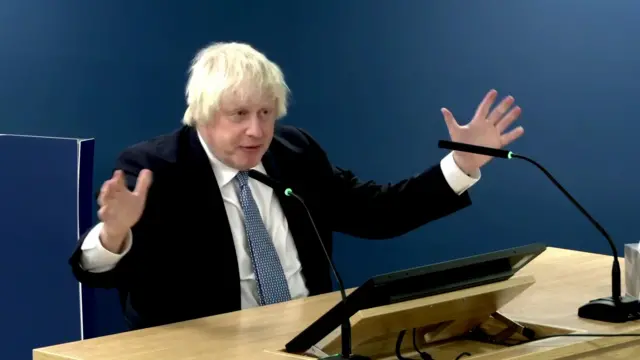 Boris Johnson is sat at a desk, his arms gesturing outward.
