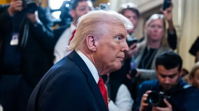Side view of Trump in a blue suit and red tie, surrounded by reporters and photographers