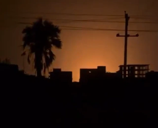 A screengrab from a video taken at night, the sky has been illuminated behind a several buildings, a palm tree and a electricity pole, which can be seen in silhouette