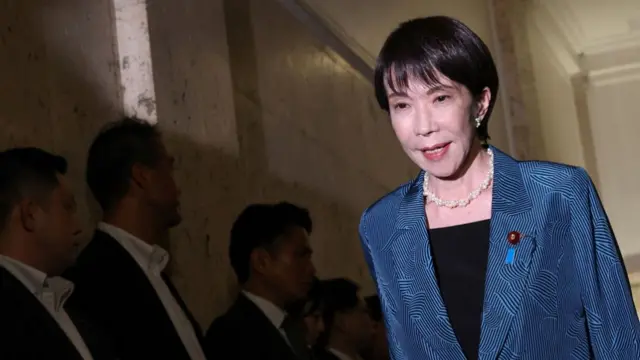 Takaichi looking to the ground as she walks along a hallway lined by men in suits. She is wearing a blue blazer and white pearl necklace.