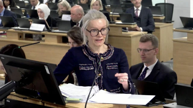Counsel Clair Dobbin asks Boris a question. She is wearing a navy cardigan and glasses, with her hair tied back.