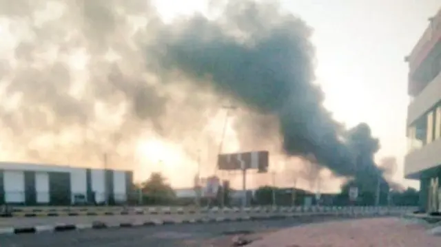 A plume of smoke rising into the sky, in the foreground is a road, a flat roofed building and a roadside billboard