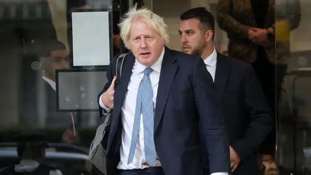 Boris Johnson in dark suit, white shirt and patterned tie hold onto a grey backpack as he walks out of the covid inquiry in Paddington, a man in a black suit following behind him