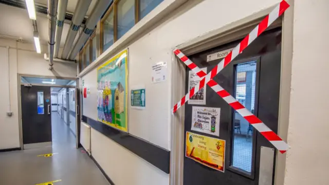 A school corridor with social distancing signs on the floor and some doors have tape over them blocking people from going in.