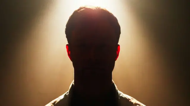 A silhouette of a man standing in front of a white light against a brown background