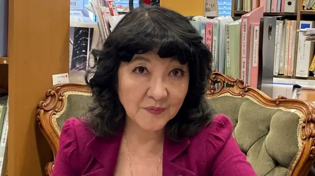 Satsuki Katayama with permed black hair smiling at the camera. She is wearing a magenta blazer and sitting on a green armchair.