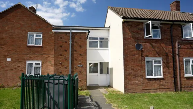 A picture of some flats on a housing estate made of red brick. There is a lawn and some bins at the front and blue sky above.