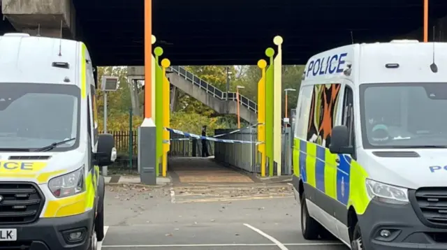 Two white police vans with blue and yellow squares on the side at the entrance to a train station with a row of yellow poles on either side