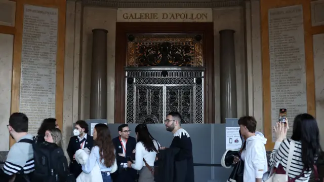 The entrance to the gallery has grey boards in front of it. People are walking past it, including one who us taking a picture on her mobile.