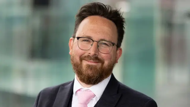 Hefin David, with dark hair a beard and glasses. He is wearing a pink tie