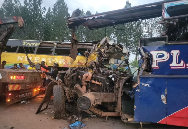 A heavily damaged bus being loaded on to a two truck, with a second heavily damaged bus in the background