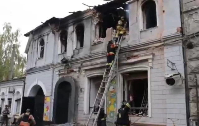 Firefighters on a ladder into a building which has a huge hole in it and burn marks, as well as blown-out widows.