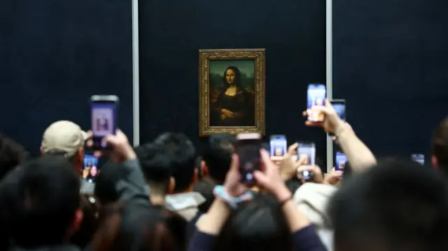 Mona Lisa painting is in the centre. Multiple people are facing it with there phones held up to take a picture of it. The wall the painting is on is navy
