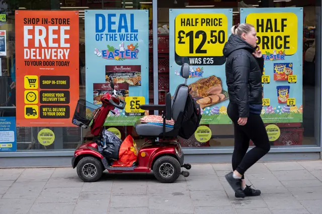 A woman walks by a grocery store, where there's a mobility scooter and signs on the store windows that show prices slashed