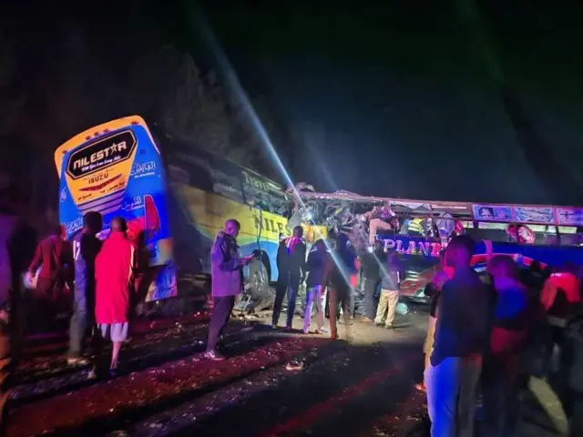A photo taken at night showing two collided buses and a crowd of people standing around them