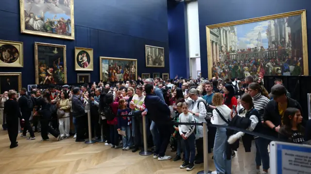 A crowd of people stand behind a corden as they talk pictures and look at the Mona Lisa, which is off camera. The walls behind them are painted navy and there are several paintings hung up on them.