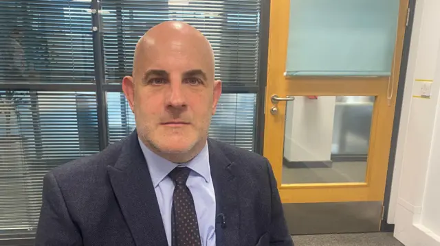 DI Neil Meade is wearing a suit and tie, looking at the camera in an office. Behind him is a door with the blinds partially closed.