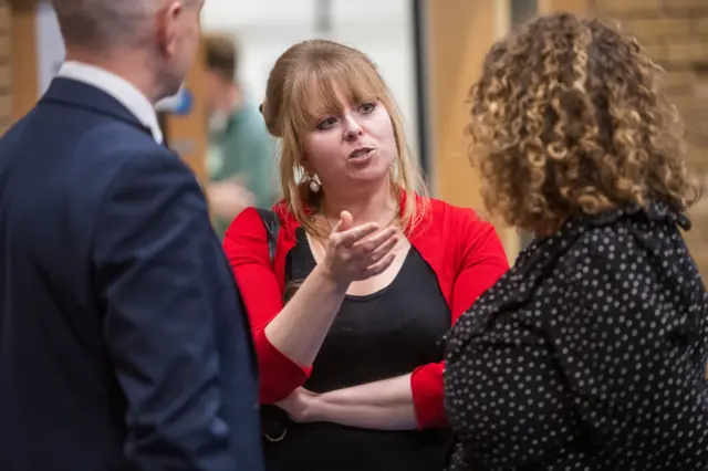 Delyth Jewell stood between two people wearing a black top and a red cardigan