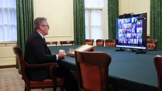 Keir Starmer sits in a yellow room at a table, in front of him is a monitor displaying a video call with lots of world leaders visible
