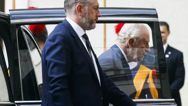 King Charles stepping out of a car, he is visible through the window which is rolled half way down.