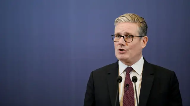 Keir Starmer in a black suit and red tie against a blue background