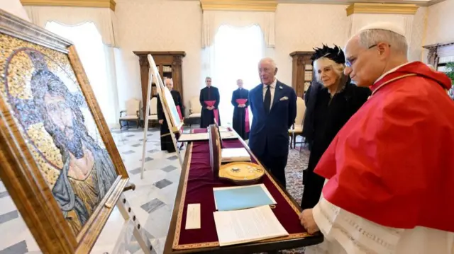 The King, Queen and Pope stand at a table where gifts have been laid out. The mosaic of Christ that has been gifted by Pope Leo is in the corner of the frame