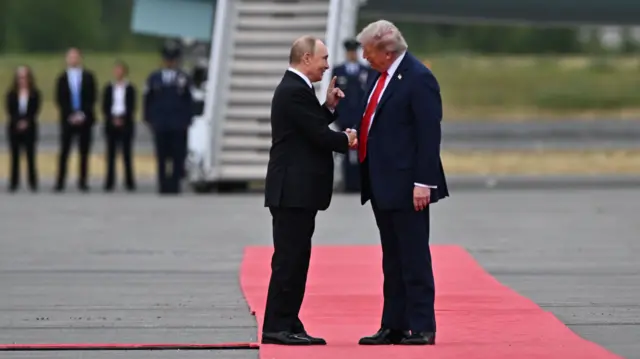 Putin and Trump stand on a red carpet on the tarmac in front of a plane, shaking hands