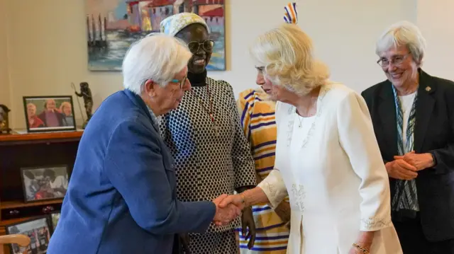Queen Camilla in a white dress shakes the hand of a women in blue