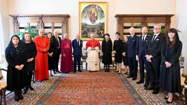 The Pope stands at the centre of a group of people. The King and Queen are on either side of him and others stand around for a photo