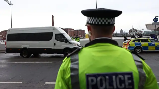 A police officer in a high vis jacket that says 'police' on it stands in front of a road. On the road there is a van which is being escorted by a police car.